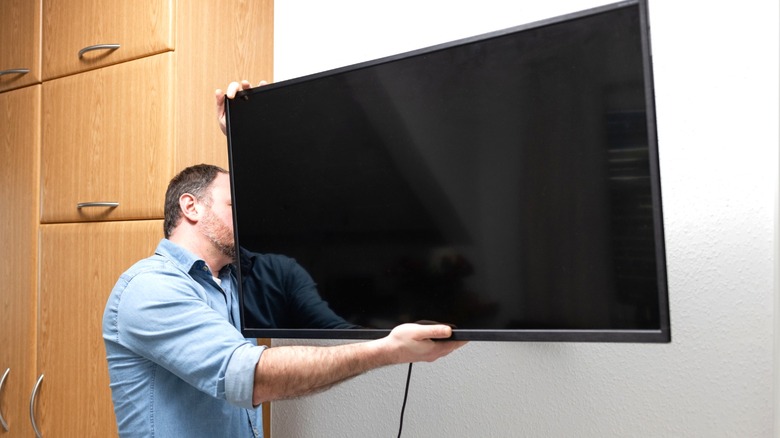 man hanging flatscreen TV on bare wall next to light wood cabinets