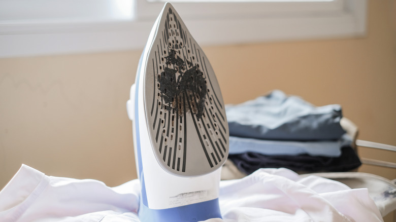 A steam iron has a dirty soleplate and a pile of clothes beside it