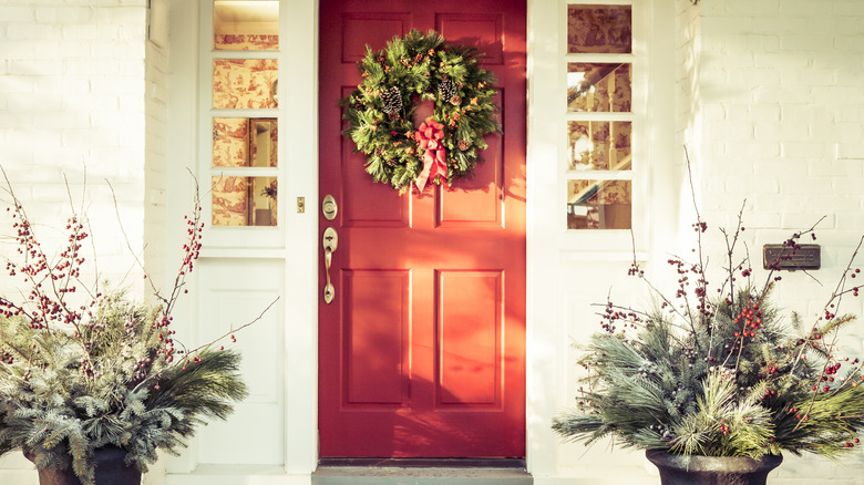 A porch with a red front door is decorated with Christmas greenery