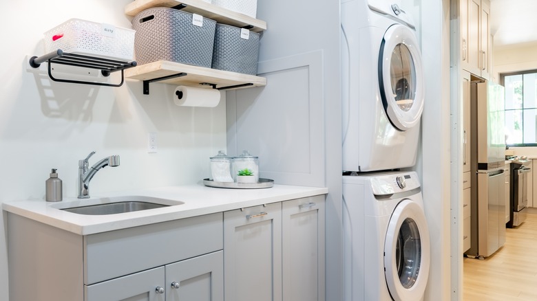 Stacked washer and dryer with sink and counter area nearby and storage from cabinets, baskets, jars, and a shelf.