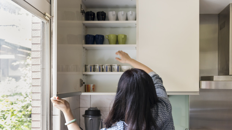 A woman reaches for one of the mugs stored on a shelf above the kitchen counter.