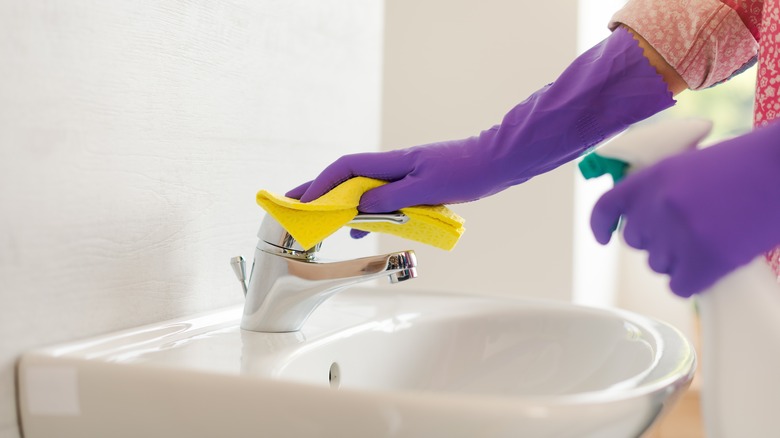 Person wearing purple gloves cleaning sink faucet with spray and yellow cloth