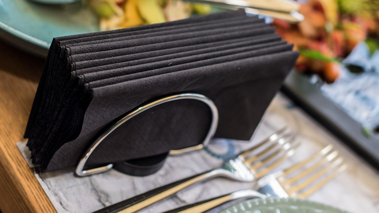 A metal napkin holder sandwiches several black napkins beside a place setting on a table.