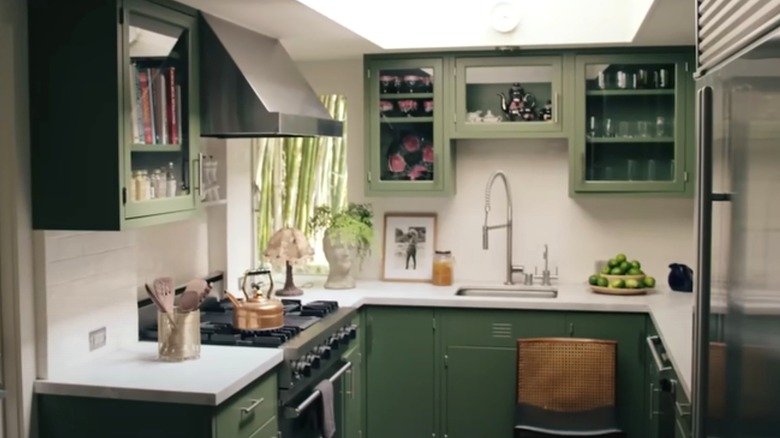 A small U-shaped kitchen features green painted cabinetry and white marble countertops along with colorful accessories