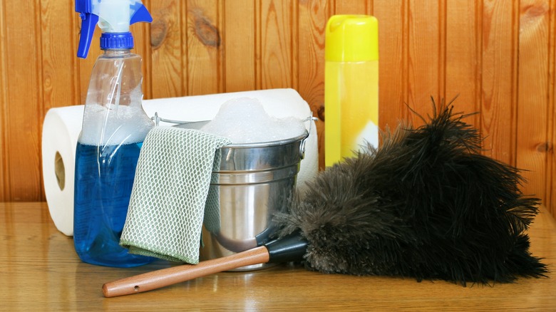 feather duster, spray bottle with cleaning liquid inside, microfiber cloth, steel bucket with soapy water and paper towel on wood background