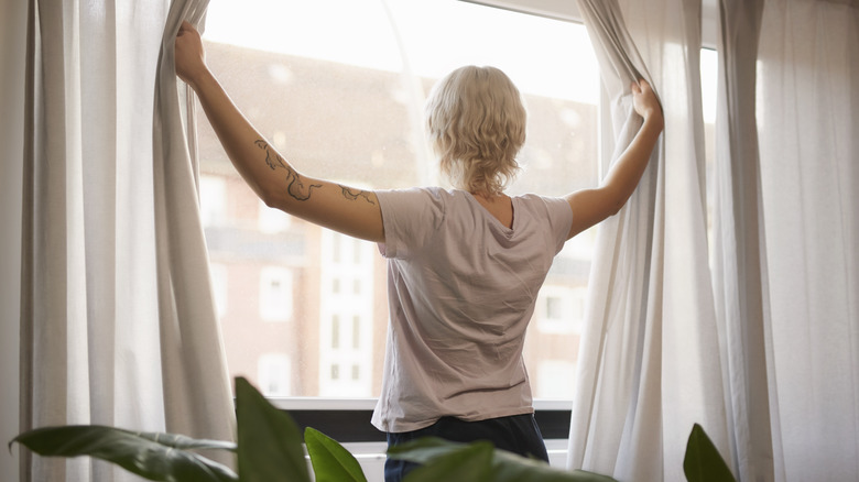 A woman pulls back curtains to look out the window