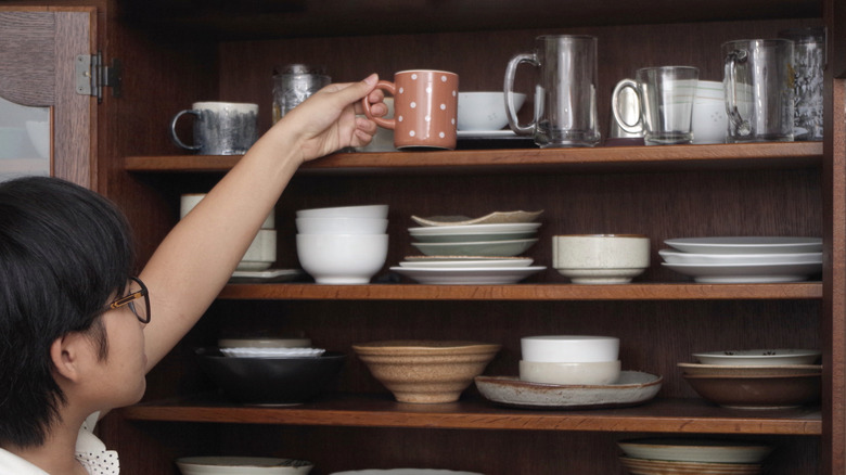 A person places a mug on a crowded cabinet shelf