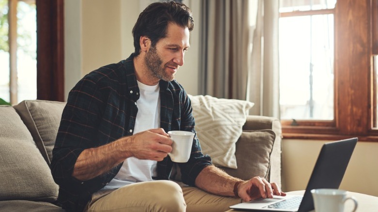 Man sitting on couch while holding mug and using laptop on coffee table