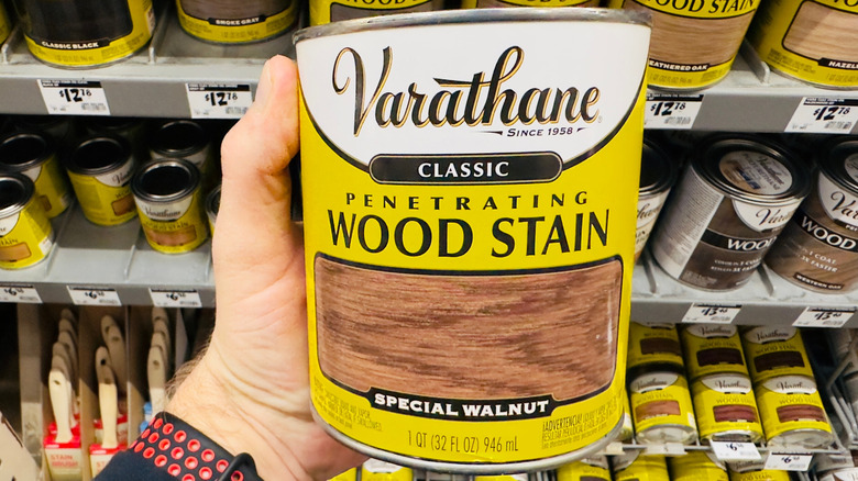 A close-up of a man's hand holding a can of wood stain in a hardware store