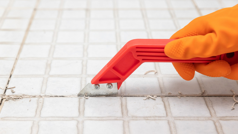 A gloved hand using a tool to scrape old grout from between tiles