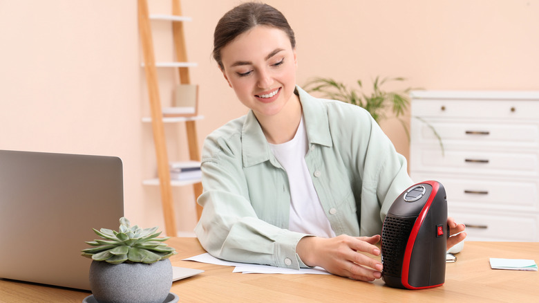 A smiling young woman adjusting a portable space heater on her desk