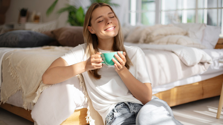 Woman sitting against her bed with a coffee mug and a smile on her face