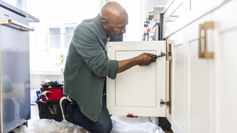 A man kneels on a kitchen floor and fixes a cabinet hinge