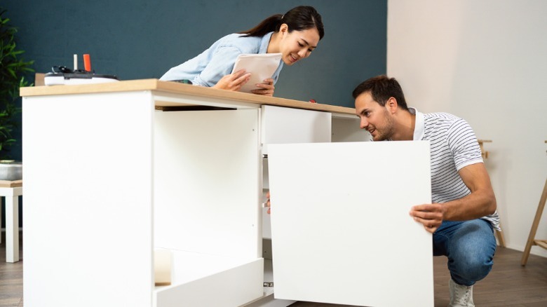 Man installing door on base cabinet while woman leans over counter watching