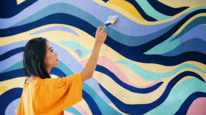 A female artist dressed in yellow painting a large mural wall.