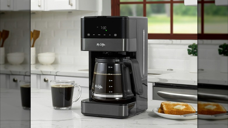 Mr. Coffee coffee maker on a white kitchen counter next to a fresh cup of coffee and a plate of toast with butter.