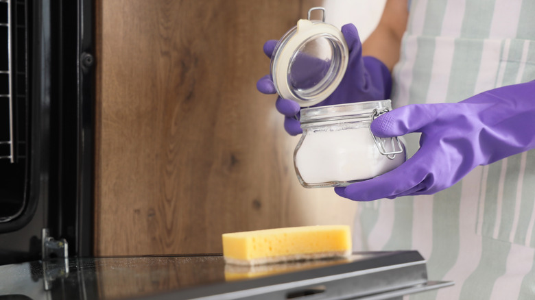 person hold jar of baking soda mixture next to open oven with sponge