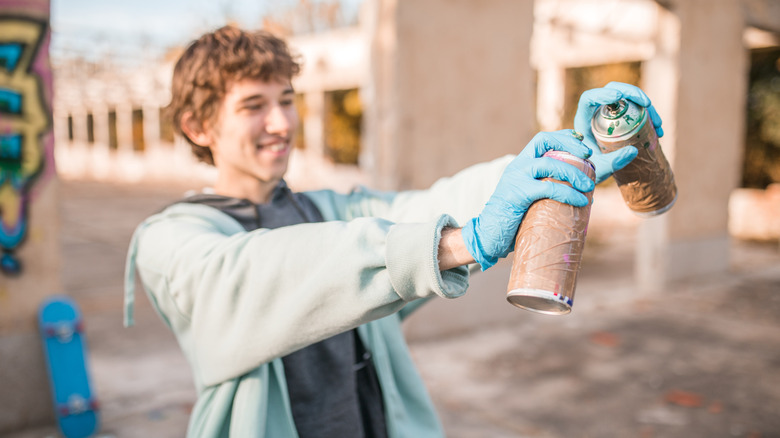 A smiling young person outdoors holding two cans of spray paint