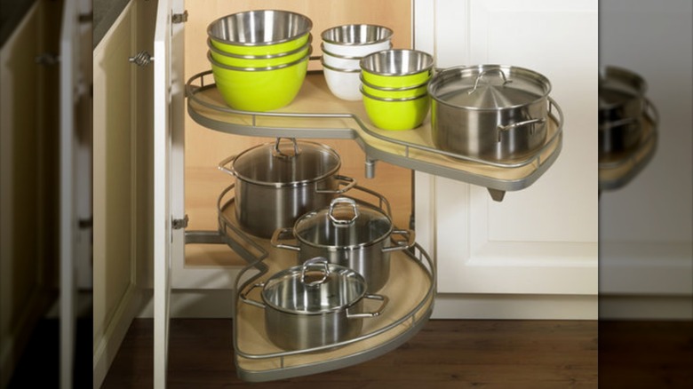 A two-tier blind shelf extends from a corner cabinet holding stainless steel cookware and bright green metal bowls