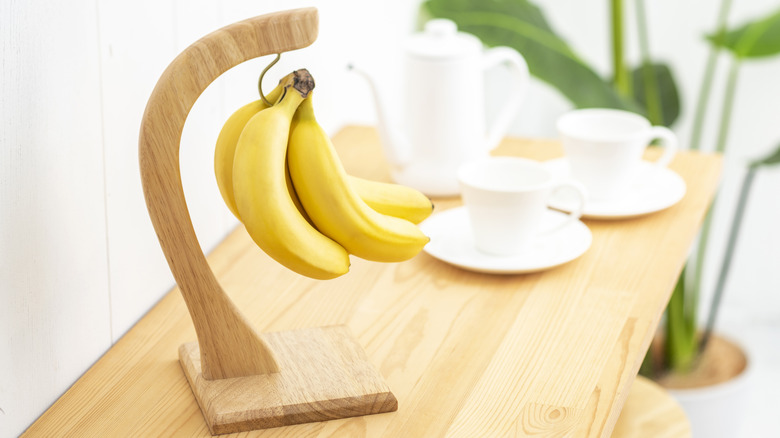 A banana hanger sites on a dining table set with a coffee pot and cups.