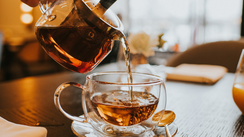 Some one pouring black tea from a kettle into a clear, glass tea cup