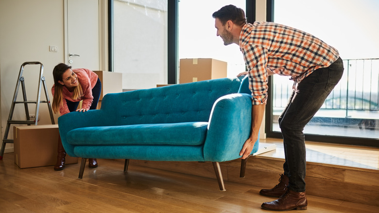 Couple moving a blue couch by carrying it over their hardwood floors