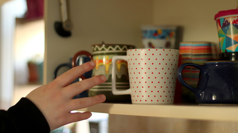 A person reaches for a mug in an open cabinet filled with mugs.