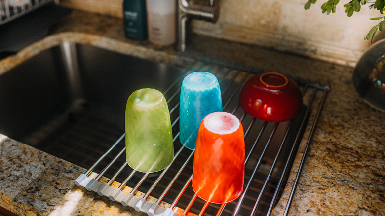 Green, blue, and orange cups drain on a roll-up drying rack next to a red bowl.