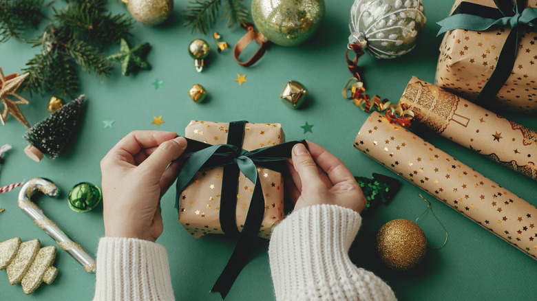 A woman wearing a white sweater tying a green ribbon on a present with wrapping supplies and accessories on a green table