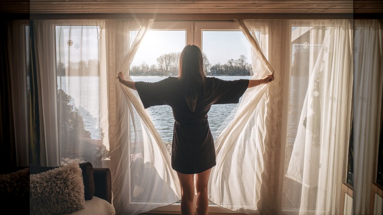 A woman opens gauzy full-length curtains to reveal a lake view