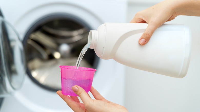 a person pouring liquid laundry detergent into a measuring cap with a washing machine in the background