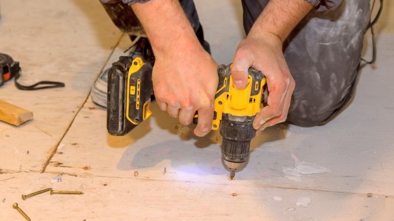 Man driving a wood screw into a plywood subfloor with a yellow cordless drill