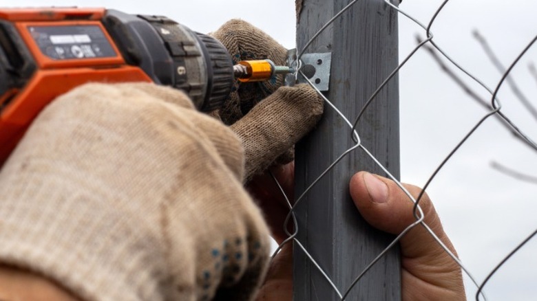 Person in work gloves drilling chain link into metal fence post extension