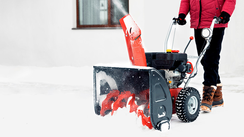 A person in a red winter jacket and black pants pushing a snow blower