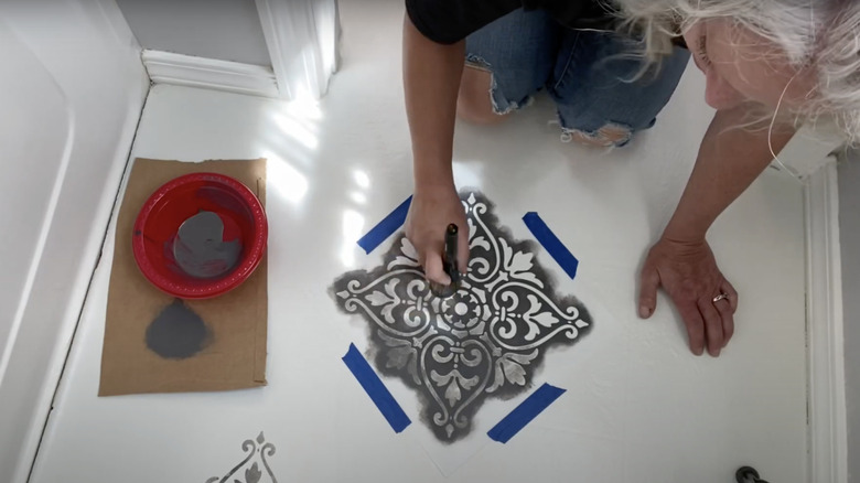 A woman stenciling white bathroom floor with gray paint