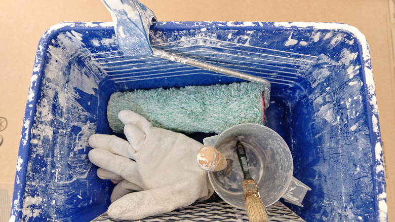 A blue paint-stained bucket containing a dried paint roller, gloves, and some painting tools.