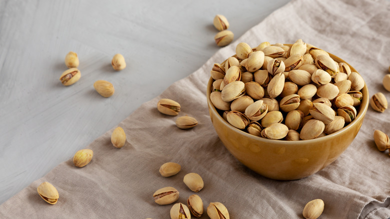 A bowl of pistachios with nuts spilling out sitting on a linen cloth.