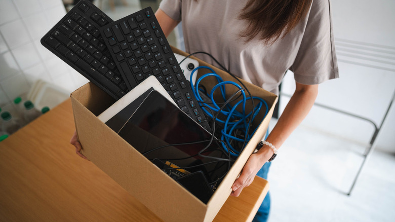 A woman holds a box of electronics including keyboards, tablets, and cables.