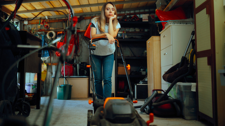 A woman leans forlornly on the handle of a lawn mower in her garage.