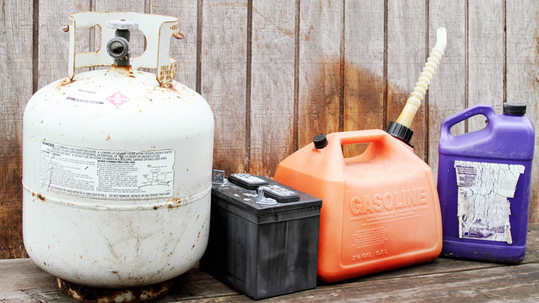 A propane tank, car battery, gasoline can, and oil bottle in a wooden-walled garage.