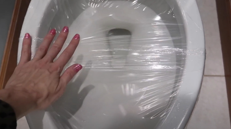 Woman's hand placing plastic wrap over a toilet bowl.