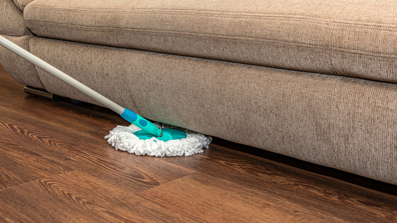 Person sliding soft mop around edge of brown fabric couch on vinyl or hardwood floor