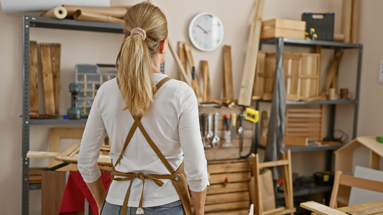 Woman in a leather apron facing wall of well-organized wood working tools