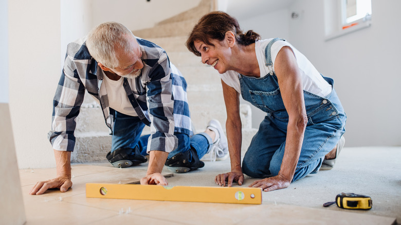 Two people installing tile flooring using a yellow tape measure and a level