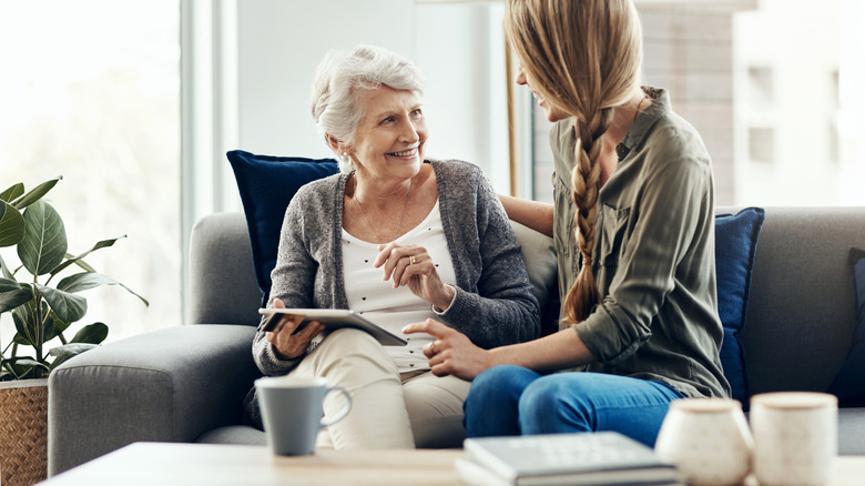 An elderly woman holding a tablet smiles at a young woman sitting next to her on a couch