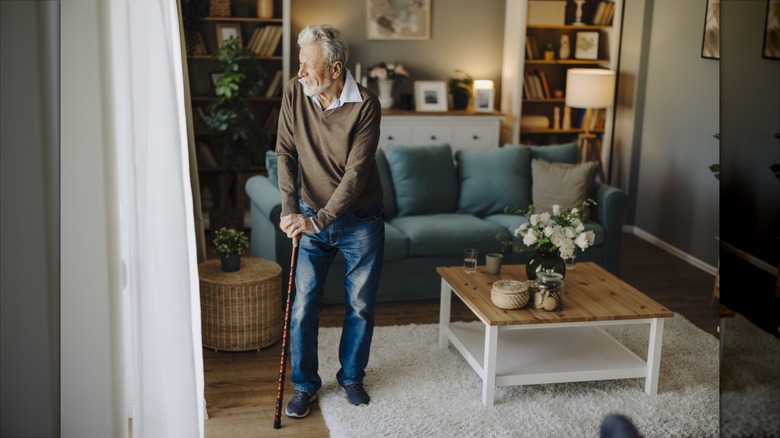 An elderly man uses a cane while looking out the window of an organized living room