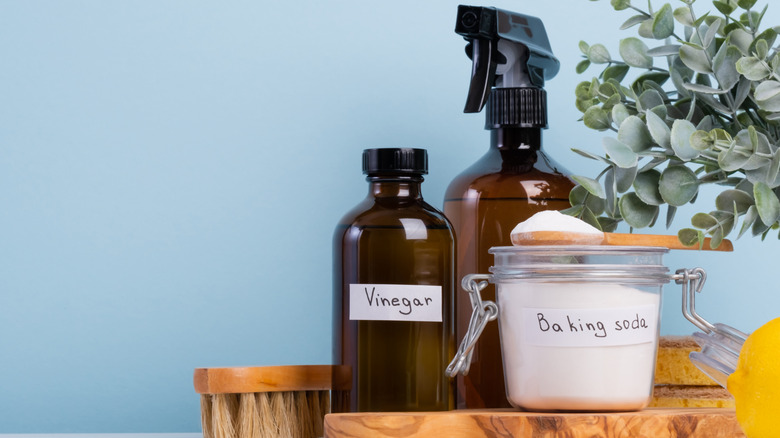 A glass container of baking soda, vinegar, and soap sit on a wood cutting board