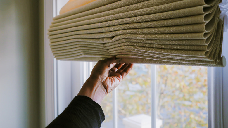 A hand holding up Roman shades fitted on a window.