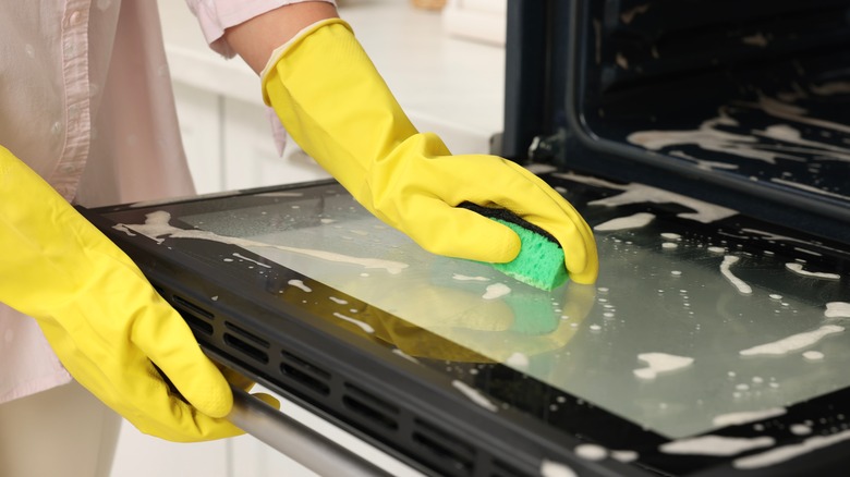 Person wearing yellow gloves cleaning a dirty oven with a sponge