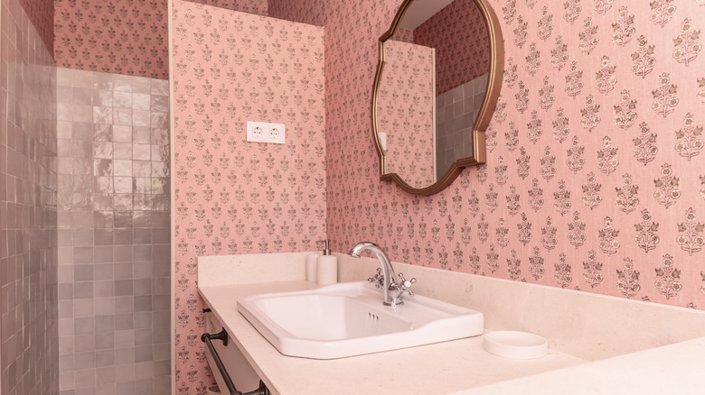 Bathroom with cream marble countertop, rose gold-framed mirror, pink wallpaper, and pink-tiled walk-in shower.